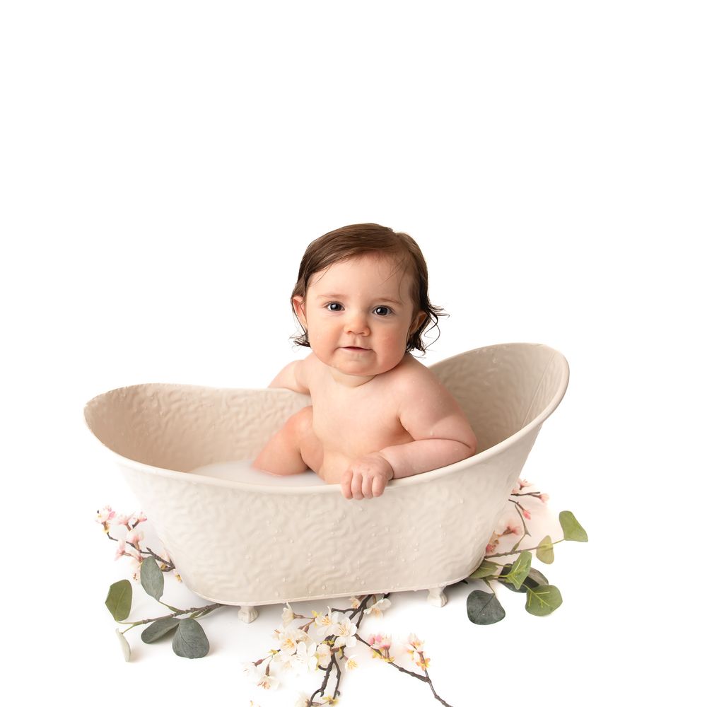 Adorable baby sitting in a cream oval bathtub prop with floral decorations for a professional photo shoot.