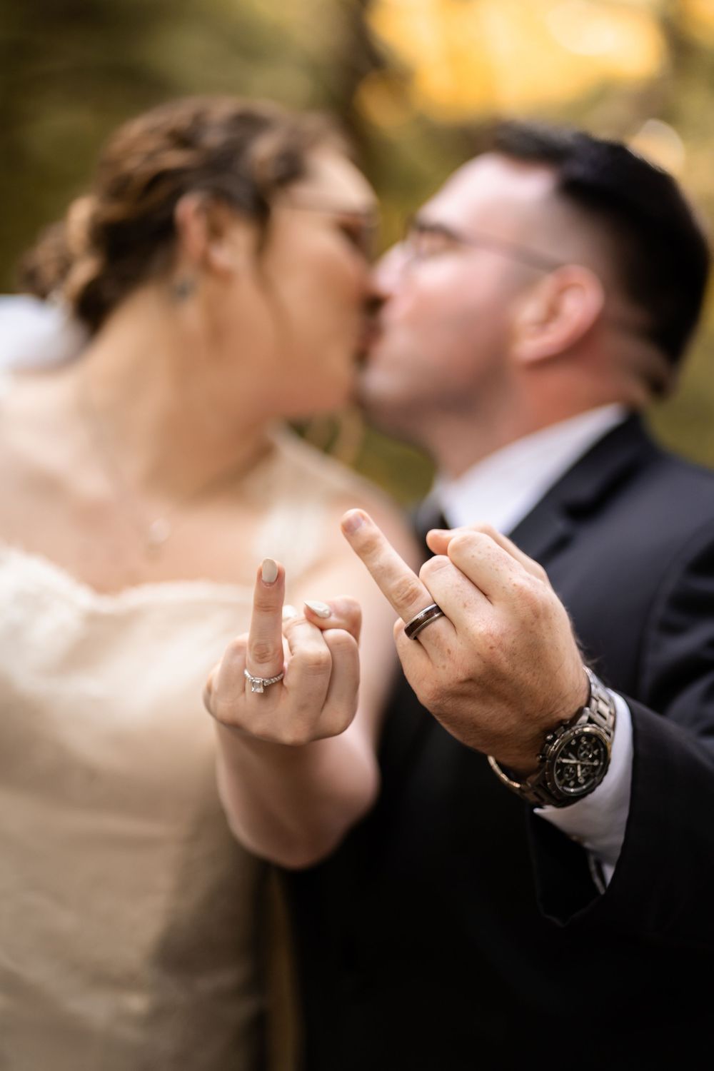 A newlywed couple shares a kiss while showing off their wedding rings and making playful hand gestures.