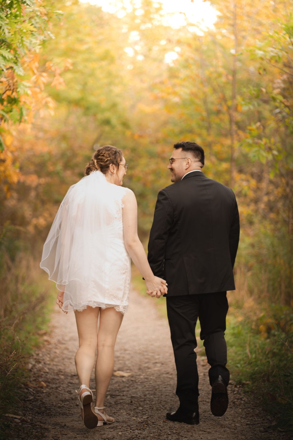 A couple in formal attire walks down a wooded path during autumn with golden foliage in the background.