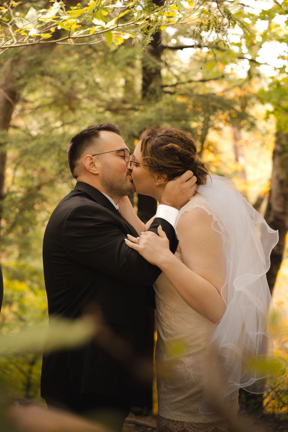 A romantic wedding couple shares intimate kisses in a sunlit outdoor setting with autumn trees.