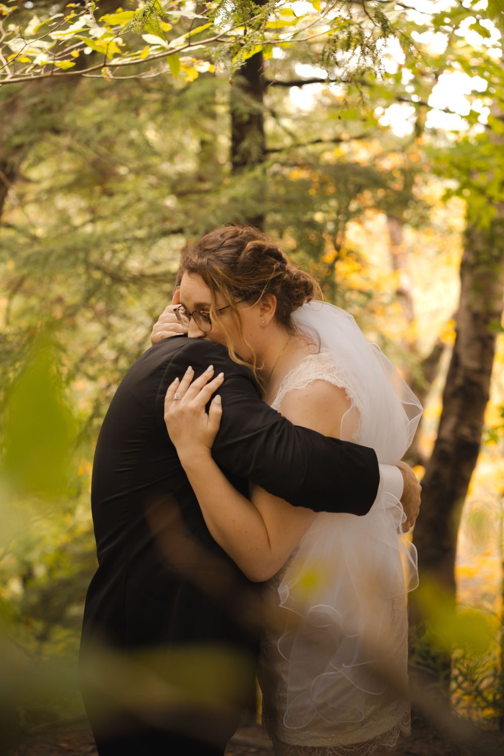 A tender embrace between a couple in wedding attire surrounded by sunlit autumn foliage in a forest setting.