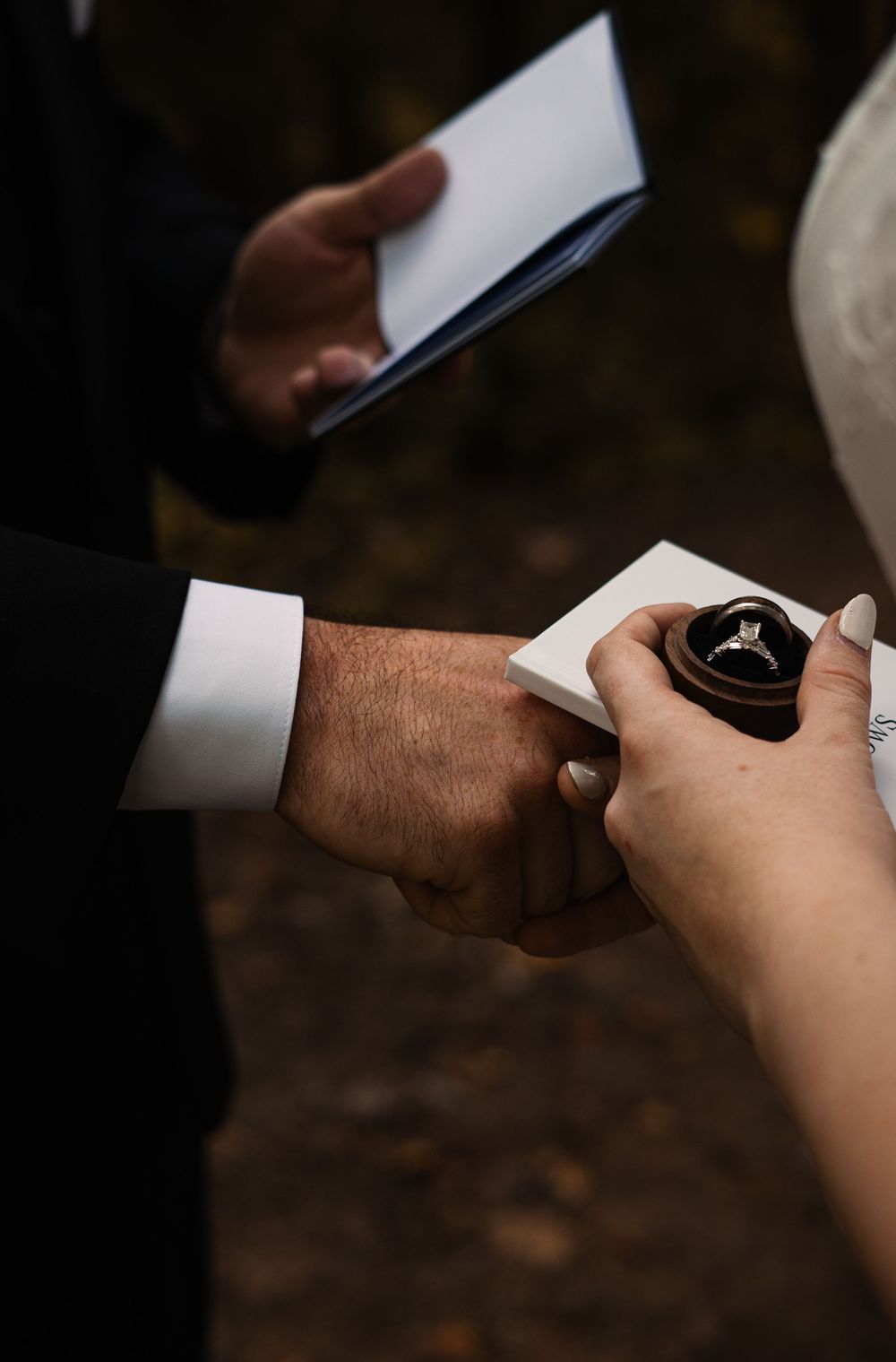 A close-up shot shows hands exchanging a wedding ring during a ceremony against a dark background.