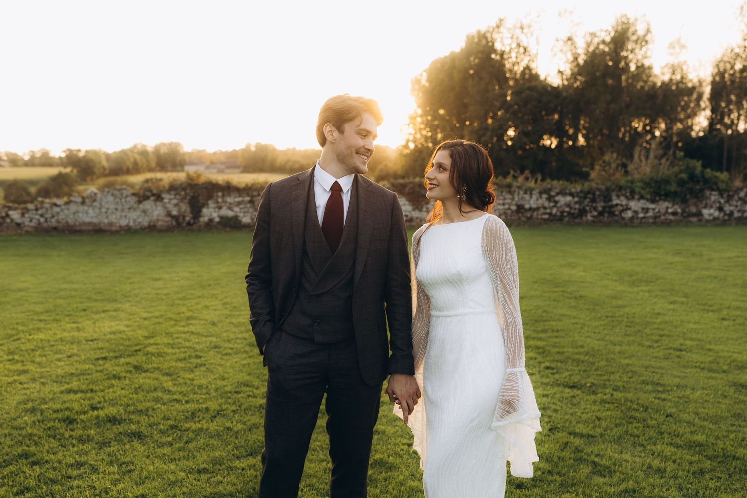 Couple in formal attire stands together on a grassy field during golden hour sunset.