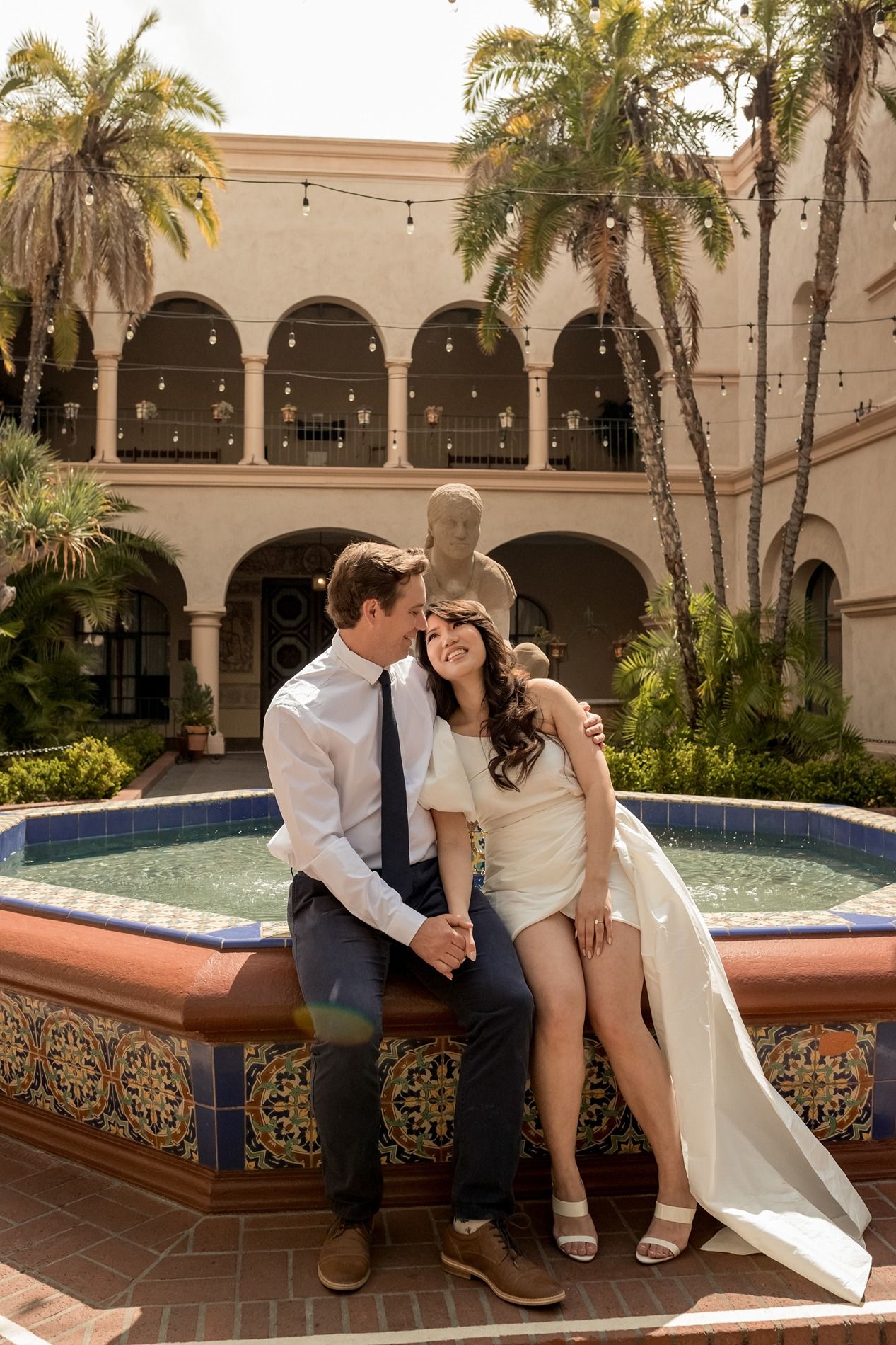 A couple sits together by an ornate Spanish-style courtyard fountain with palm trees and arched architecture in the background.