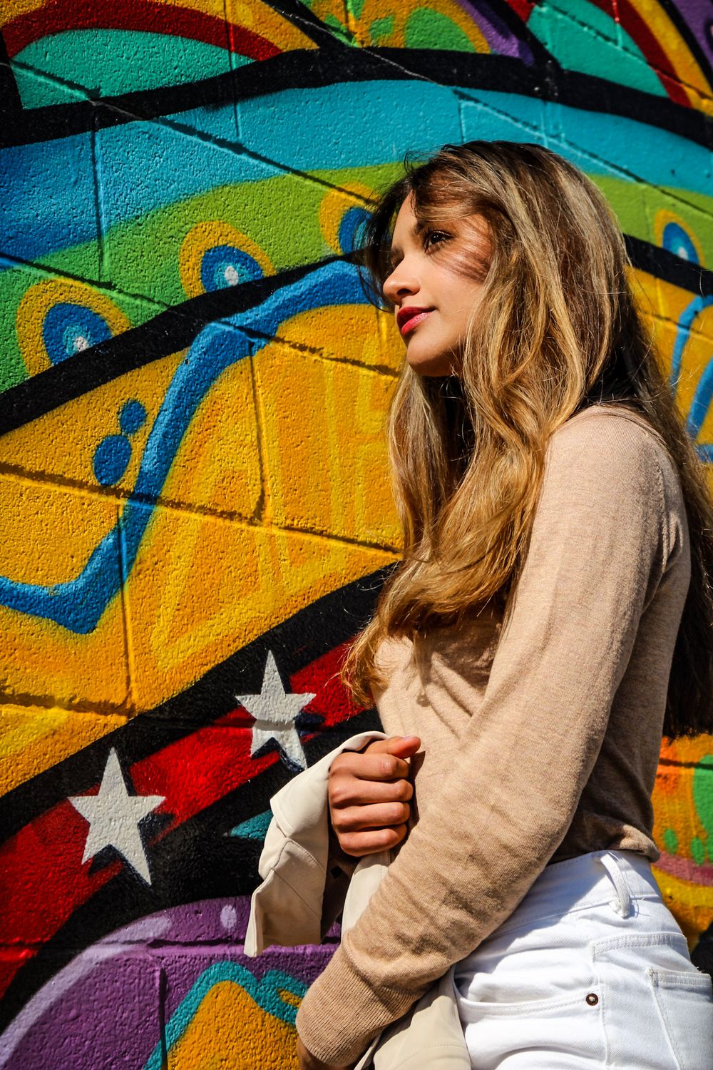 Person in beige sweater poses against vibrant graffiti wall with yellow, purple and red colors and star designs.