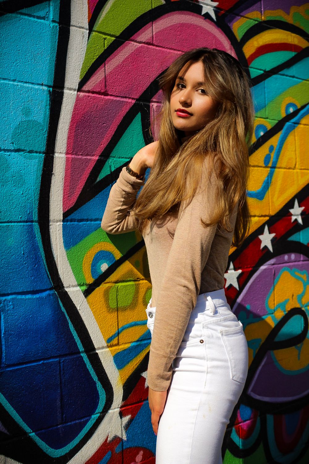 Fashion model poses against vibrant graffiti wall wearing beige sweater and white pants in urban photoshoot series.