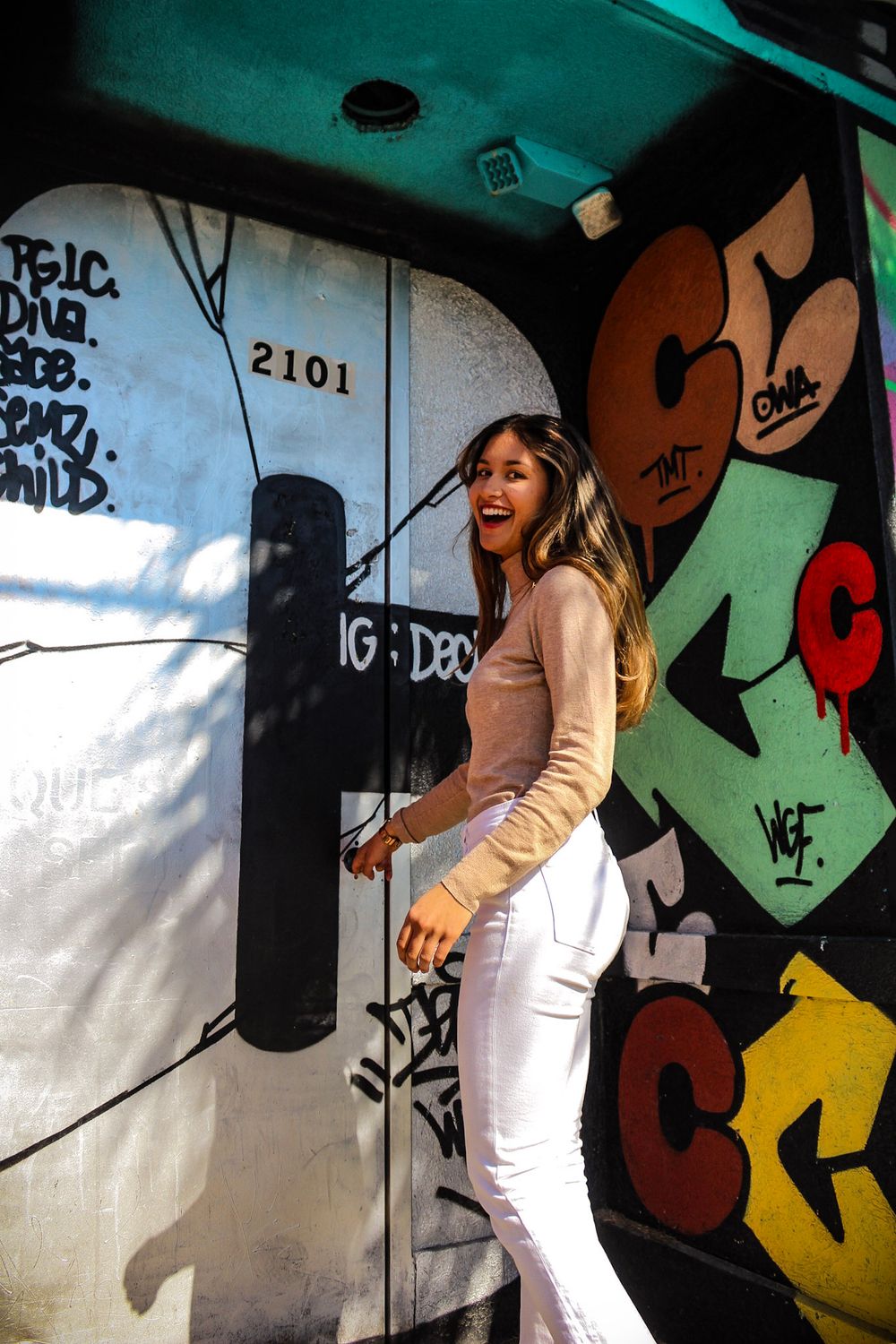 Person in white outfit dances against colorful graffiti wall with street art in urban setting.