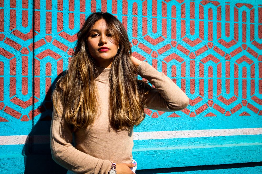 A person in a beige sweater poses against a bright turquoise and black geometric patterned background.