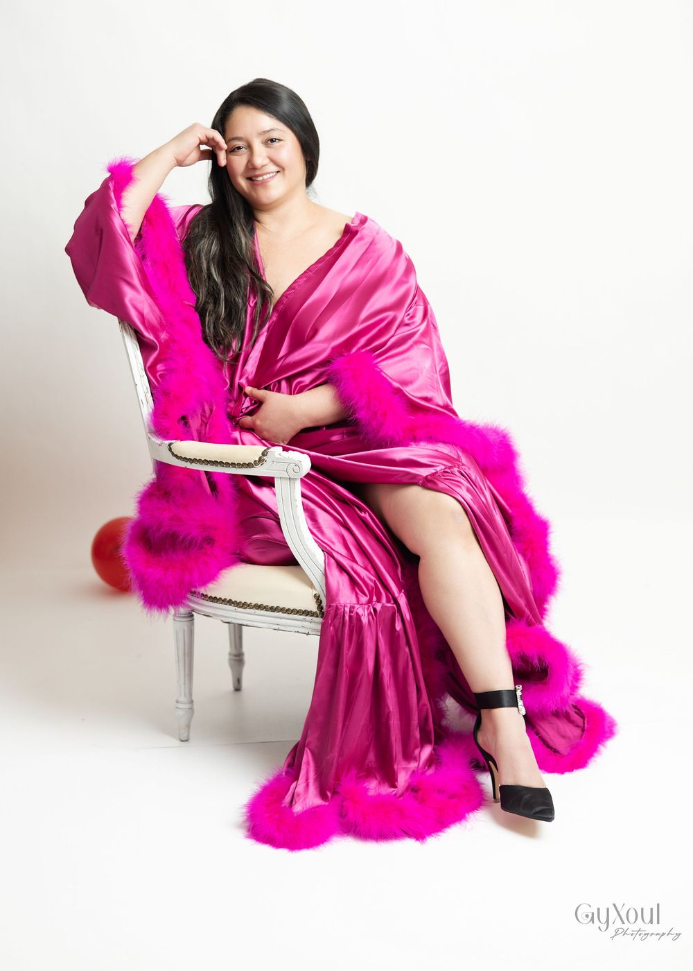 A glamorous boudoir photoshoot featuring a hot pink silk robe and matching feather boa on an elegant white chair.
