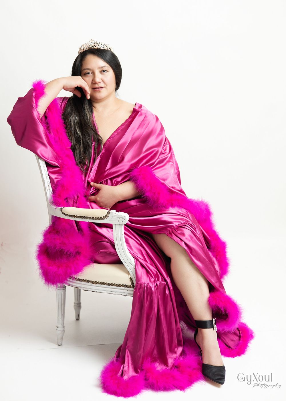 Glamorous hot pink silk gown with feather trim detail and tiara on model posing elegantly in white chair.