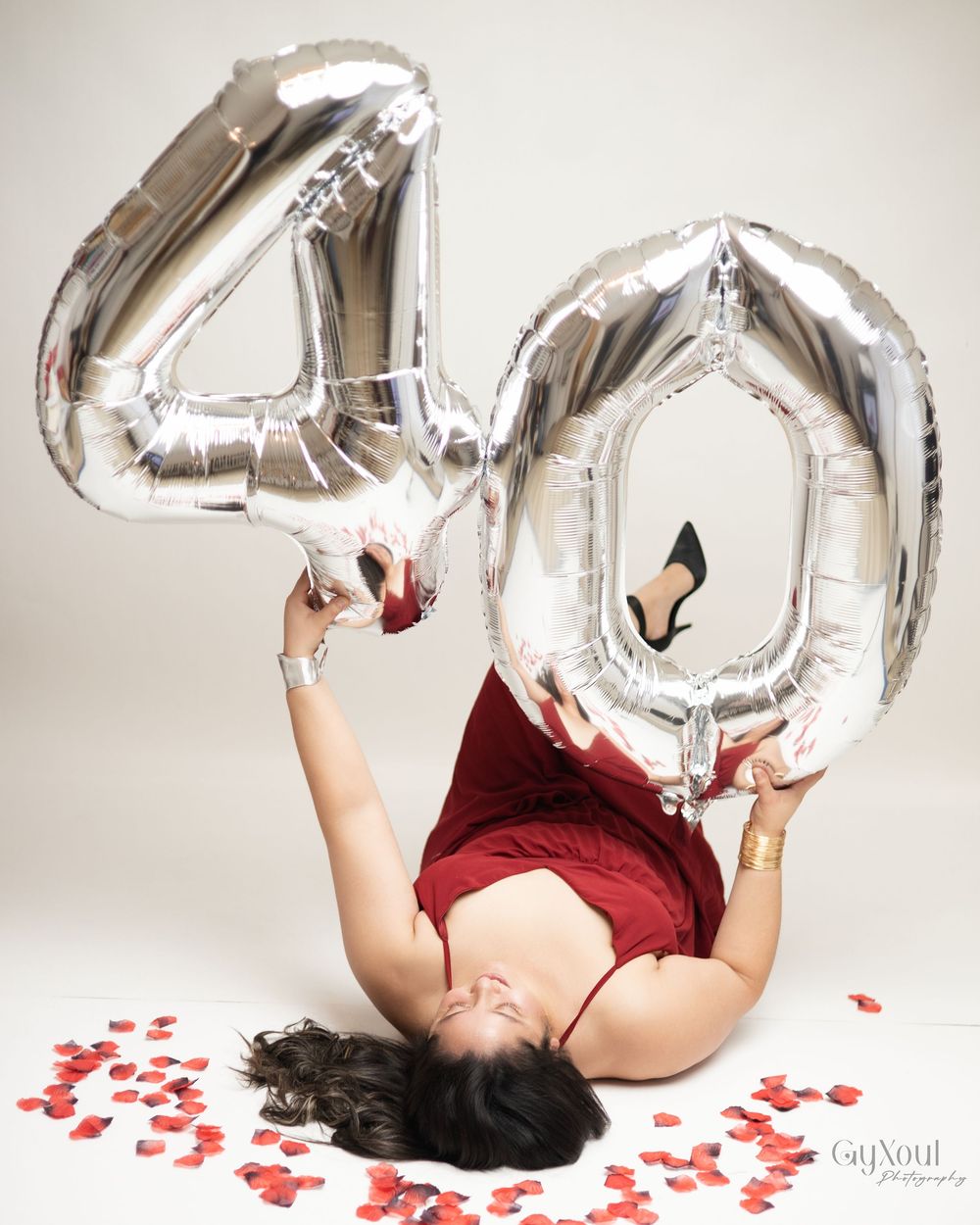 Person lying on floor with silver number balloons and scattered red heart confetti in Valentine's Day themed photoshoot.
