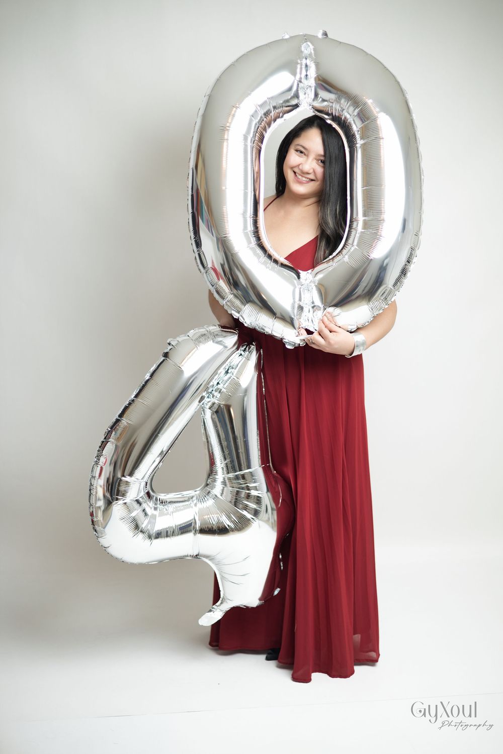 A figure in a burgundy dress holds large silver metallic number balloons against a white studio backdrop.