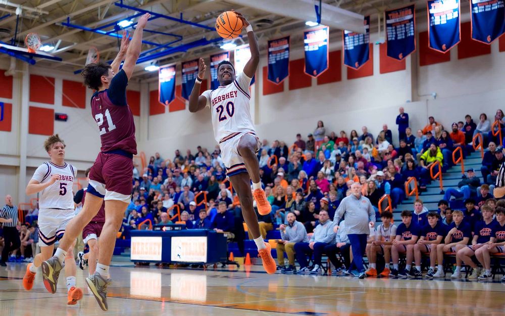 Hershey Trojans player attempts a basket with a full student section cheering behind by SportsFox Photography, Hershey PA.