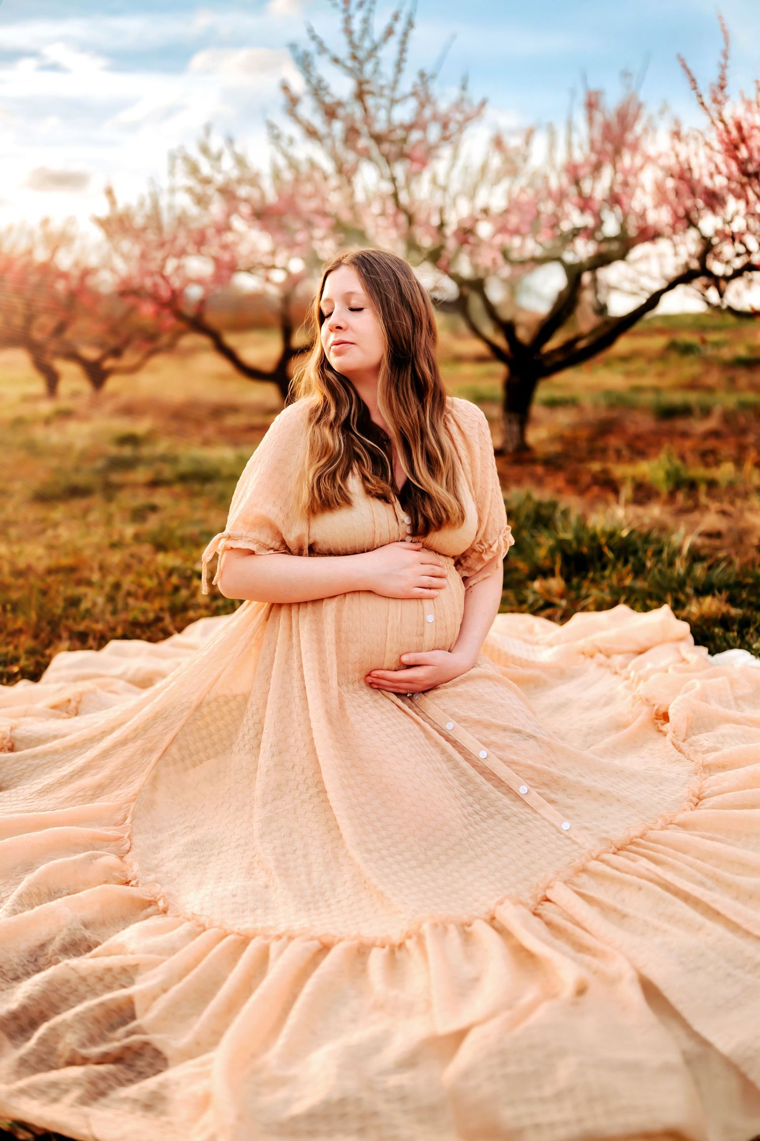 A flowing peach gown spreads out in a circular shape beneath blooming spring trees in an orchard at sunset.