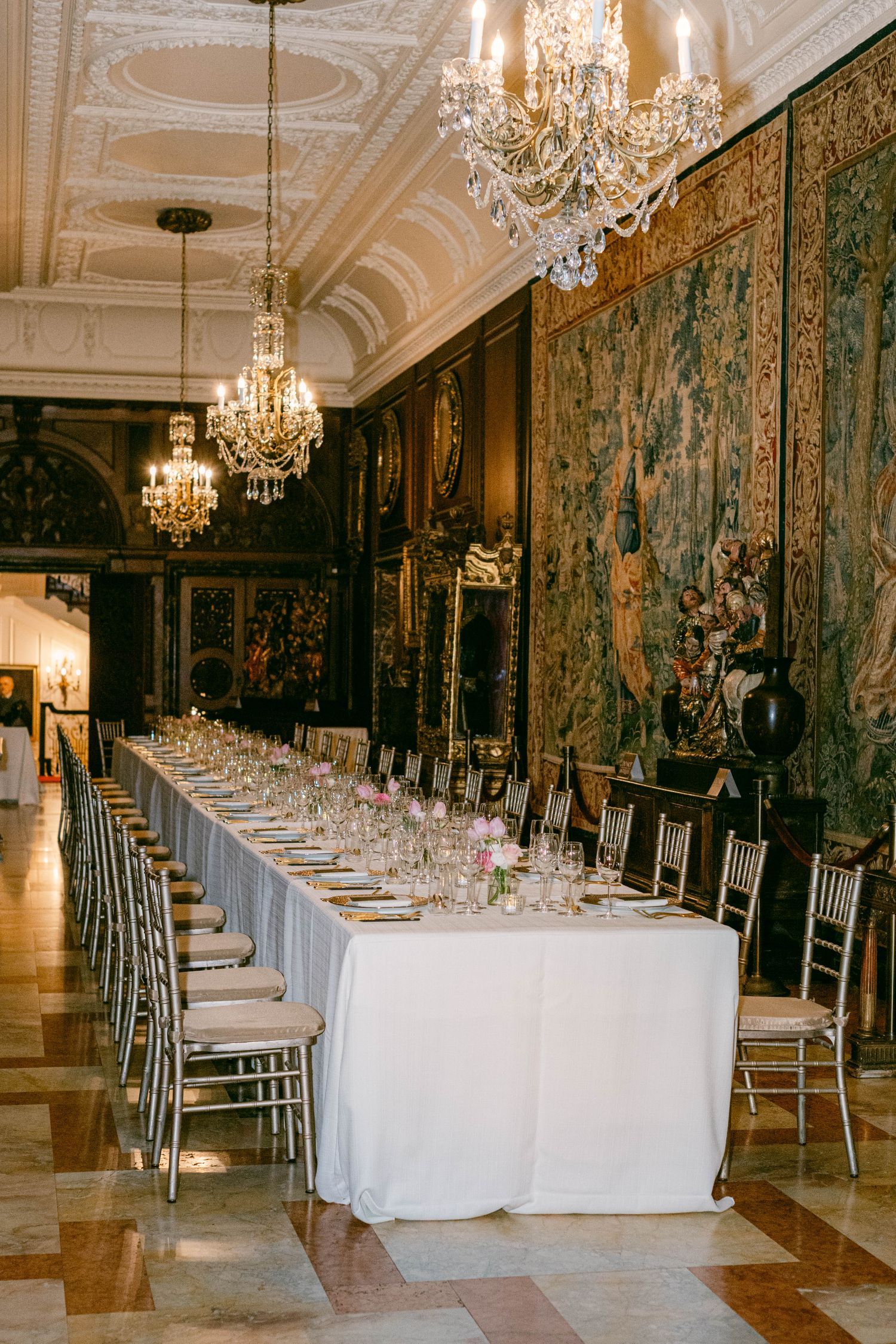 Elegant banquet hall with crystal chandeliers, ornate tapestries, and a long white-clothed dining table set for a formal event.