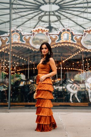 A woman in a layered rust-colored dress poses in front of a vintage carousel with twinkling lights at dusk.
