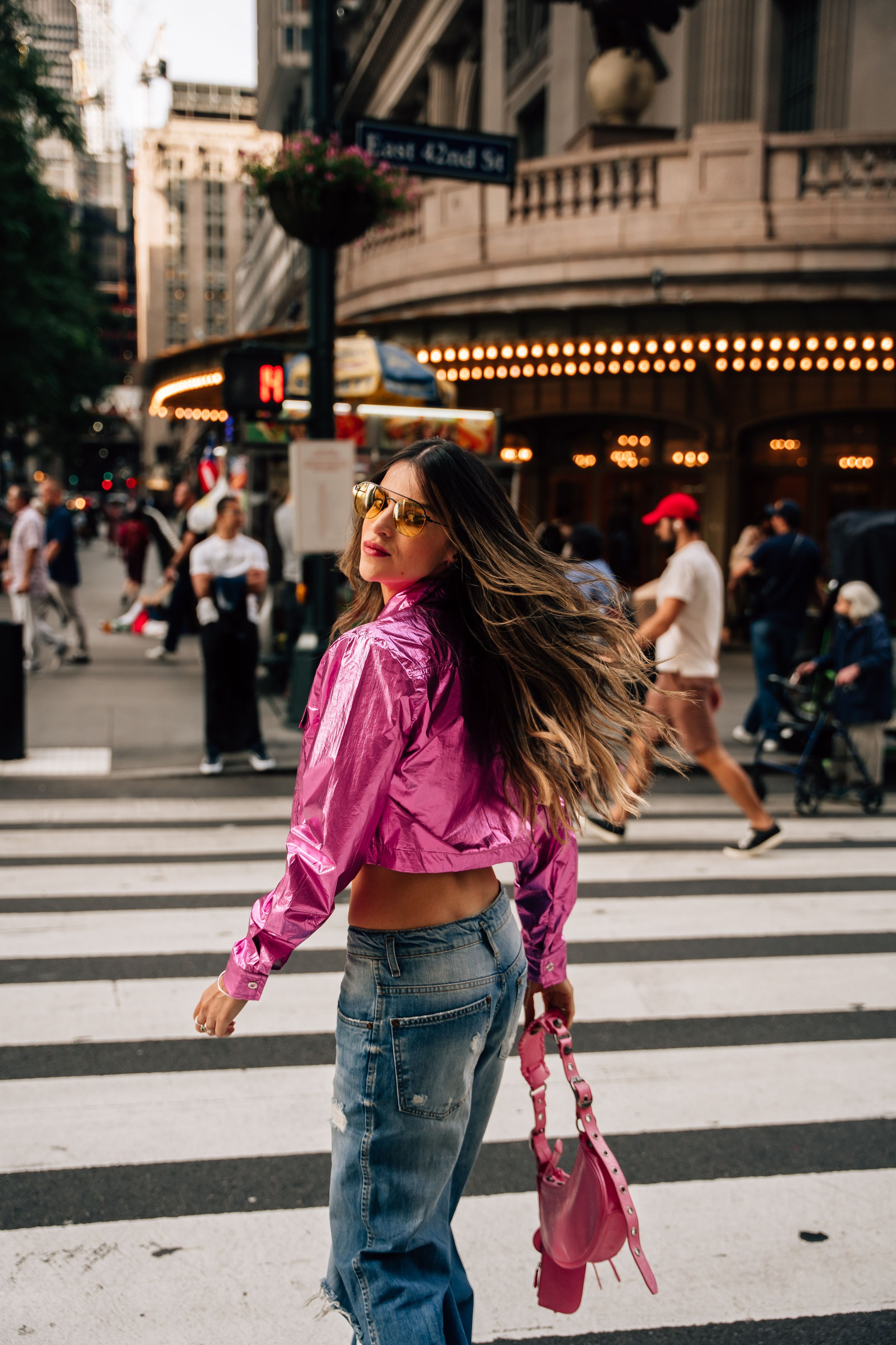 Fashionable woman in pink crop top and jeans twirling on city crosswalk with illuminated marquee lights in background.