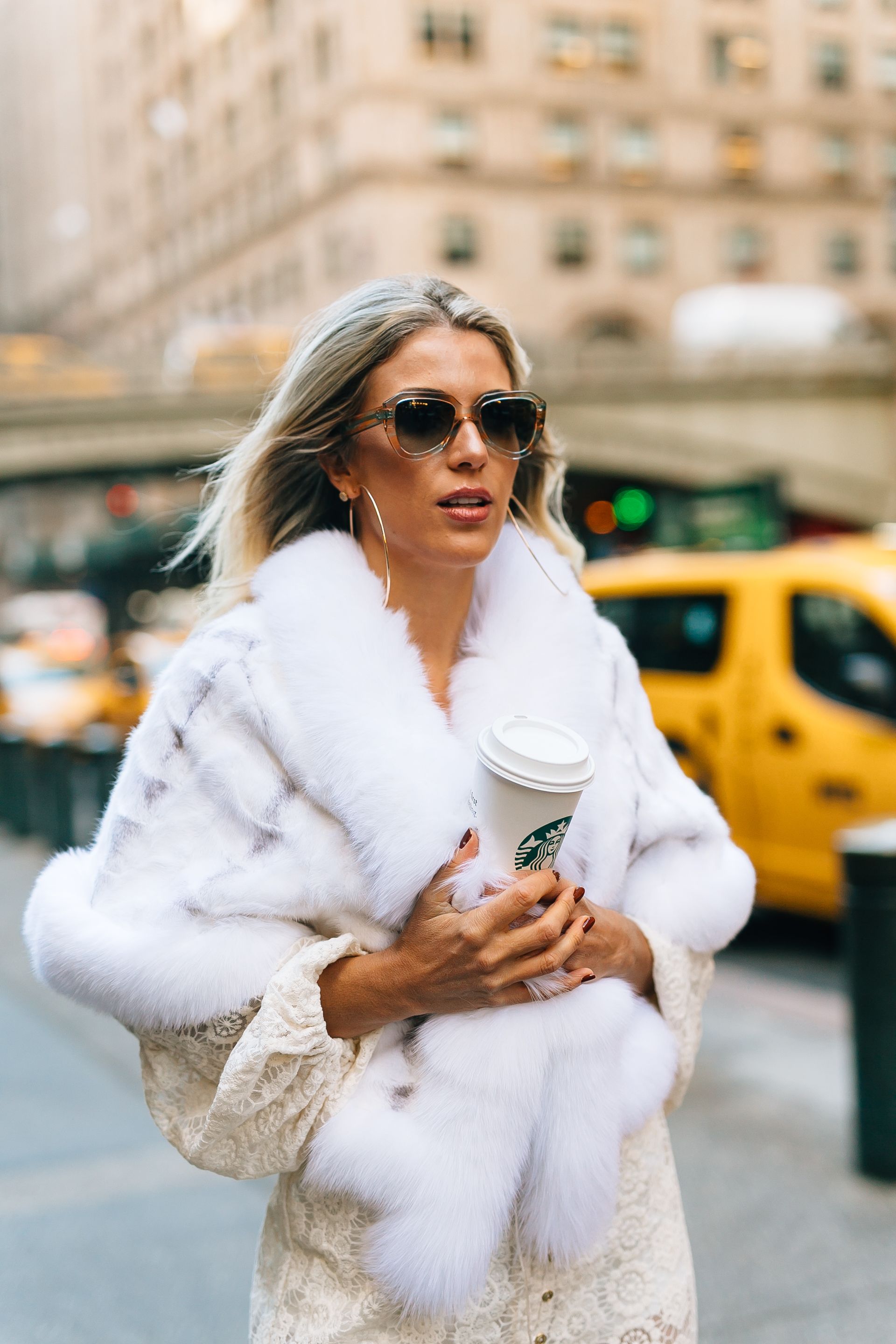 A person in a luxurious white fur coat and sunglasses walks through a city street with yellow taxis in the background.