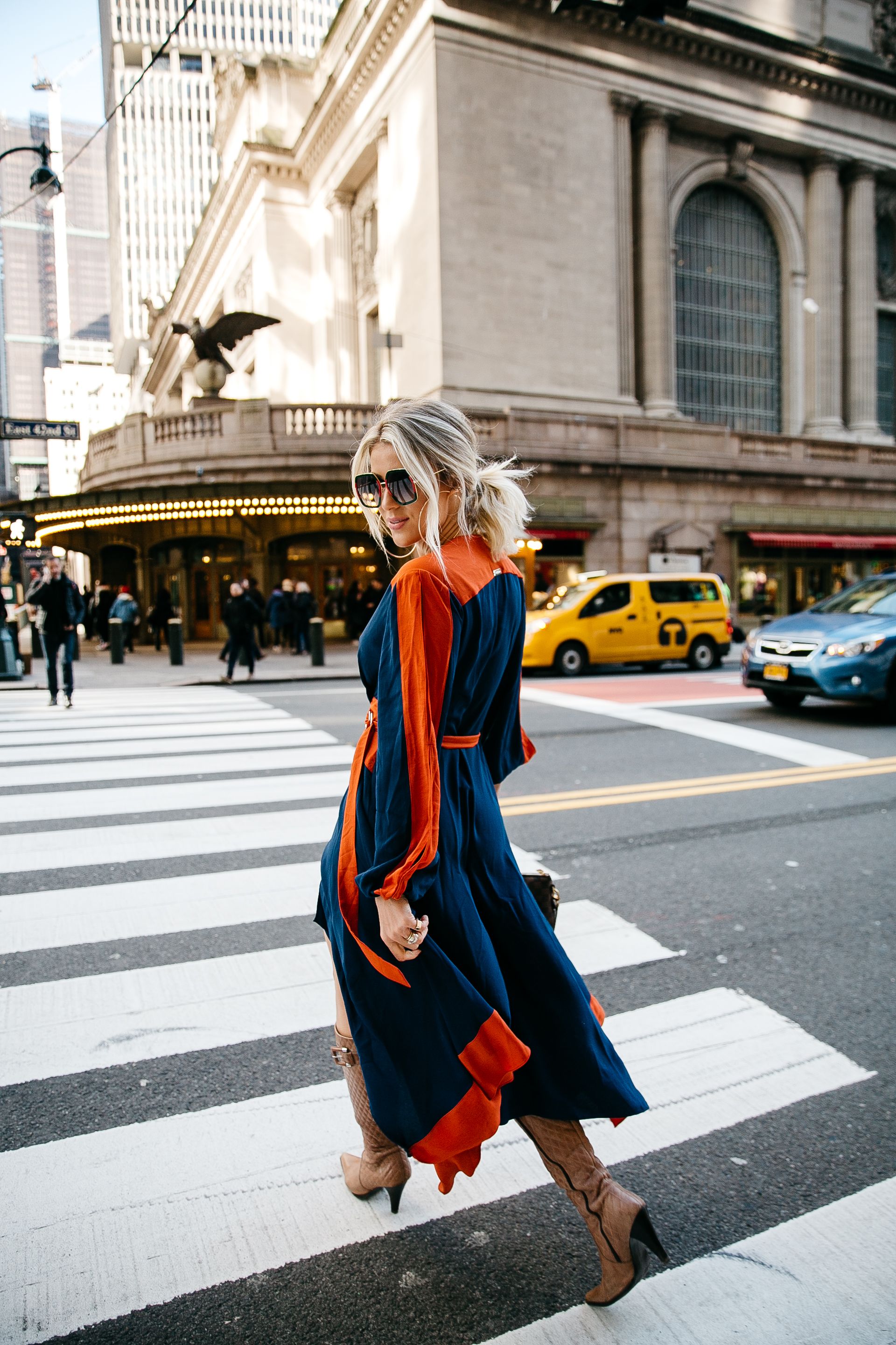 Stylish figure in navy and orange dress crosses busy New York City street near Grand Central Terminal with yellow taxis.