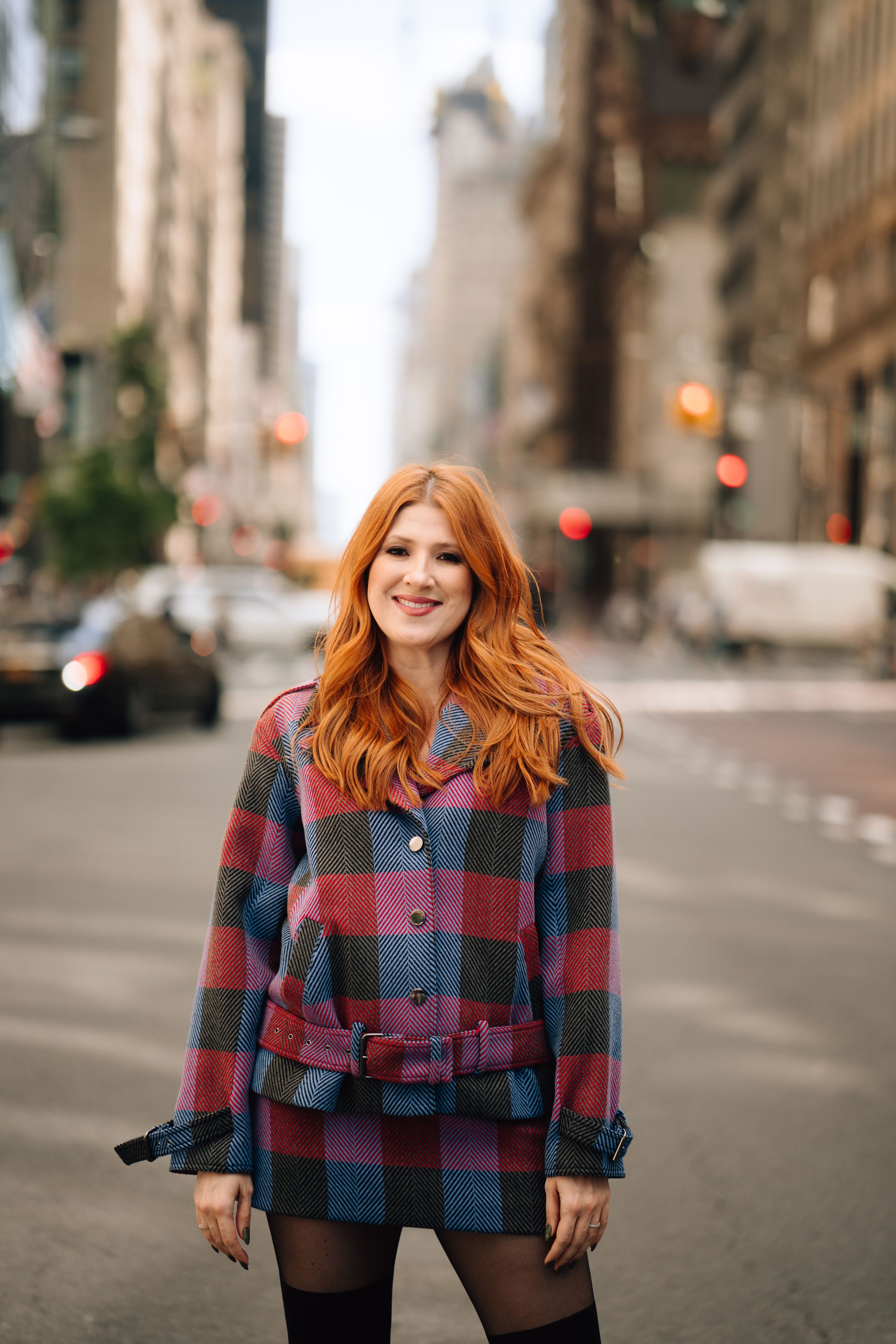 A person wearing a plaid coat smiles on a city street with tall buildings in the background.