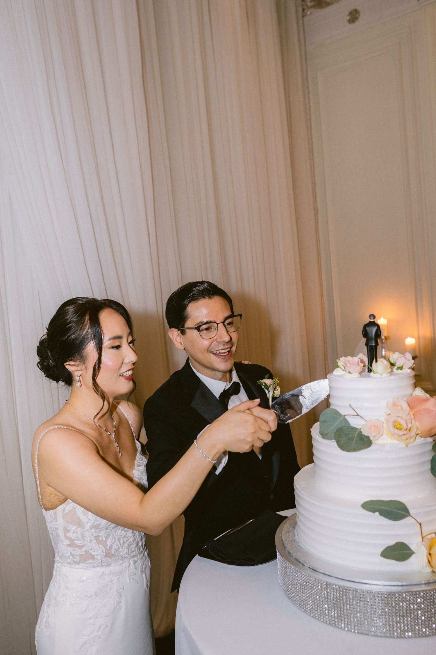 A couple in formal wedding attire cuts their white tiered wedding cake together at their reception.