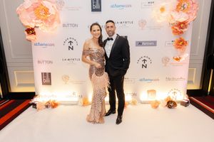 A couple poses at a formal event in front of a white backdrop with peach floral decorations and sponsor logos.