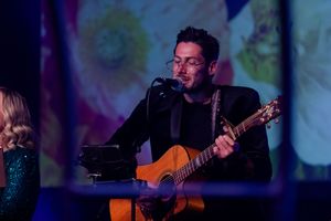 Musicians perform on stage with acoustic guitars under moody blue lighting at an evening event.
