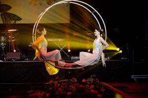 Two performers gracefully balance on a circular aerial hoop under dramatic yellow stage lighting.