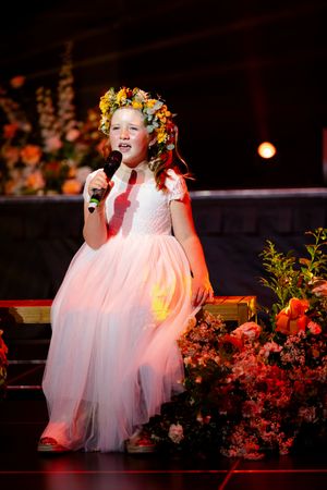 Young performers in white dresses with floral crowns sing on stage during a show.