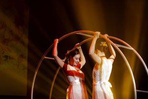 Performers in white dresses dance with illuminated hoops on a darkened stage.
