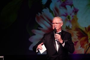A speaker holds a microphone on stage with colorful floral projections in the background.
