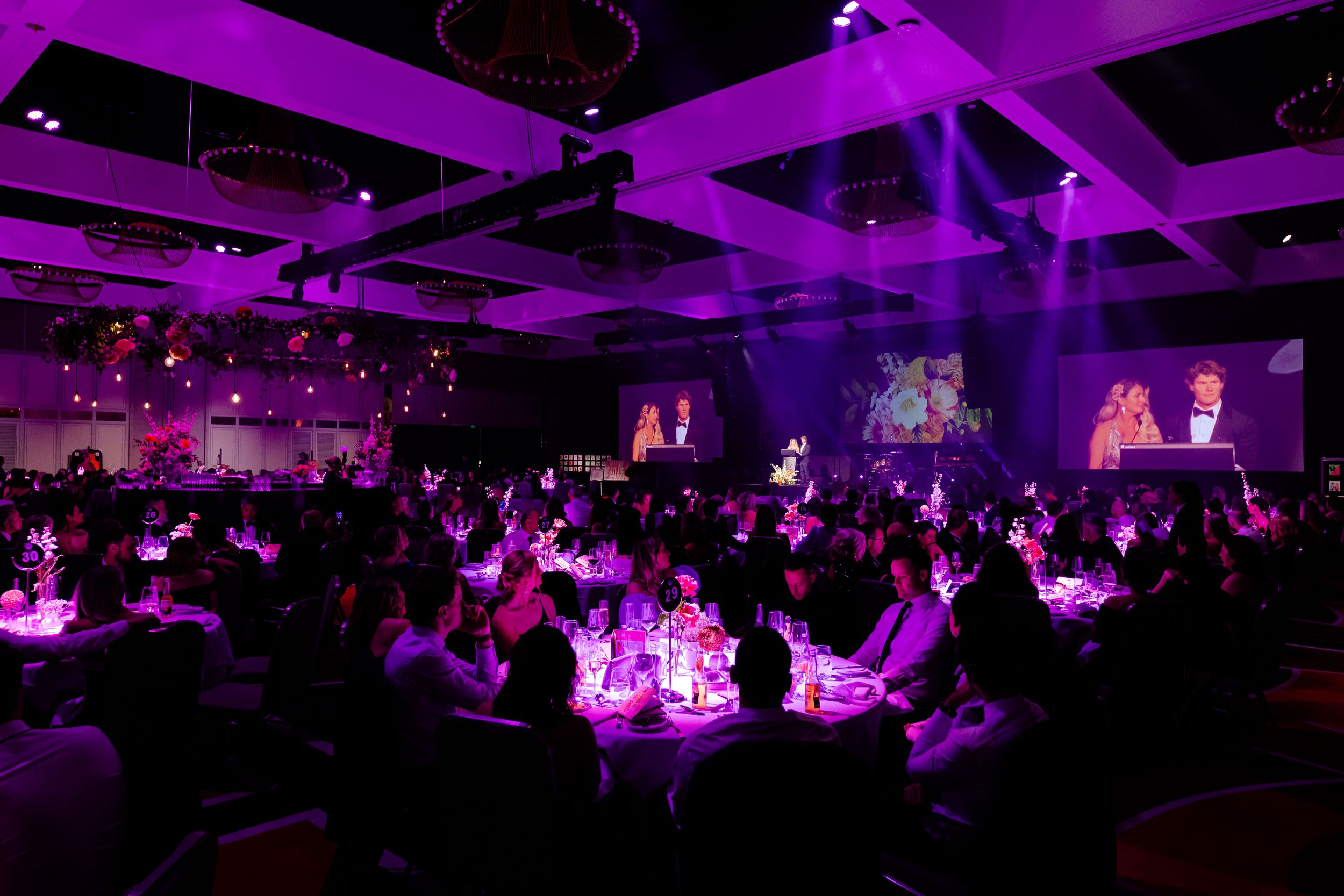 Elegant ballroom event space illuminated in purple lighting with guests seated at round tables for a formal gathering.