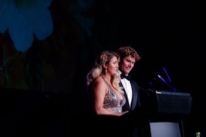 A formal couple stands at a podium during an elegant evening event with dramatic lighting.