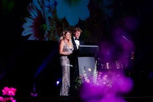 Two people speaking at a podium during an elegant evening event with colorful floral projections in background.