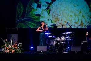 A performer plays drums on a dimly lit stage with blue lighting and floral arrangements.