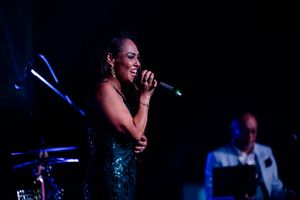 A performer in a sparkling dress sings on stage with dramatic lighting and a band member visible in background.