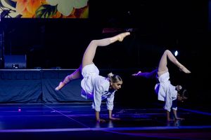 Two dancers perform synchronized handstands in white outfits on an illuminated blue stage.