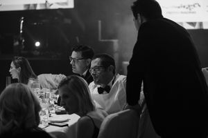 Dimly lit event space with silhouettes of attendees seated at tables during a presentation.