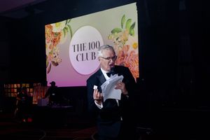 A speaker presents on stage at The 100 Club with colorful floral graphics displayed on the backdrop screen.