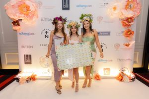 Three people wearing floral crowns and light-colored dresses pose with a decorative sign at an elegant event.