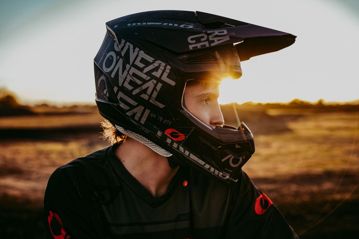 A sequence of motocross riders in black helmets silhouetted against a golden sunset during a race.