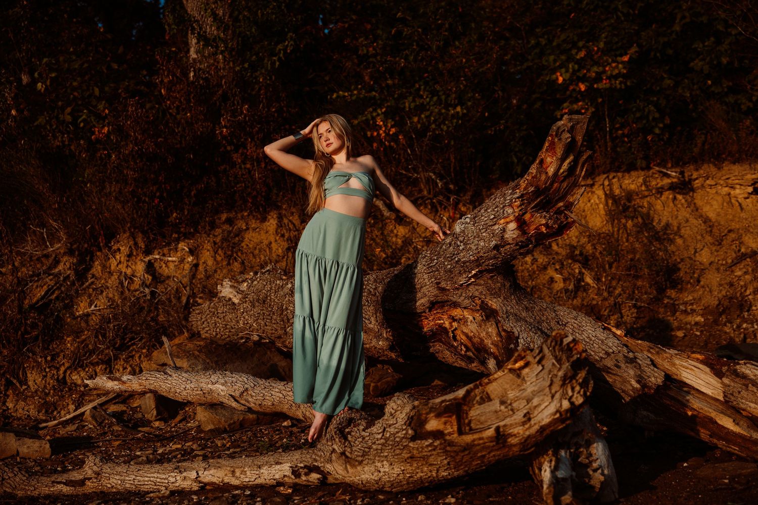 A person in a turquoise dress poses gracefully near fallen trees and rocks in a dramatic natural setting.