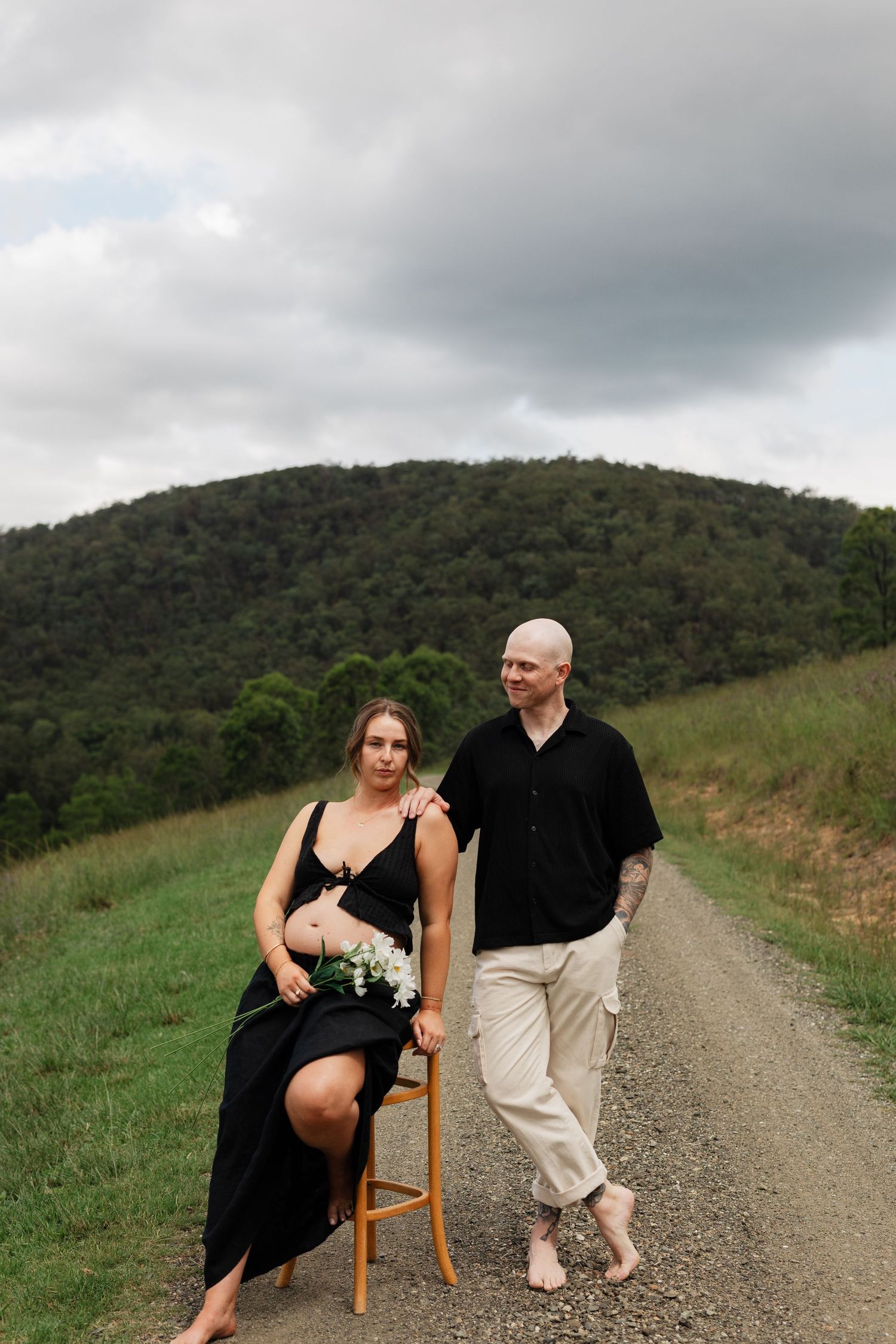 A couple poses playfully on a wooden chair along a scenic dirt road with green hills in the background.