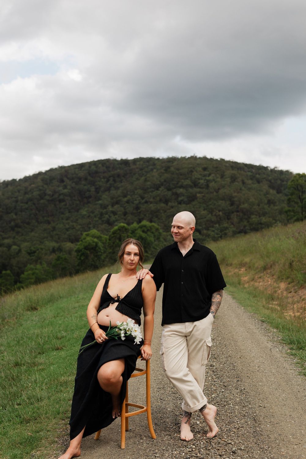 A couple poses playfully on a wooden chair along a scenic dirt road with green hills in the background.
