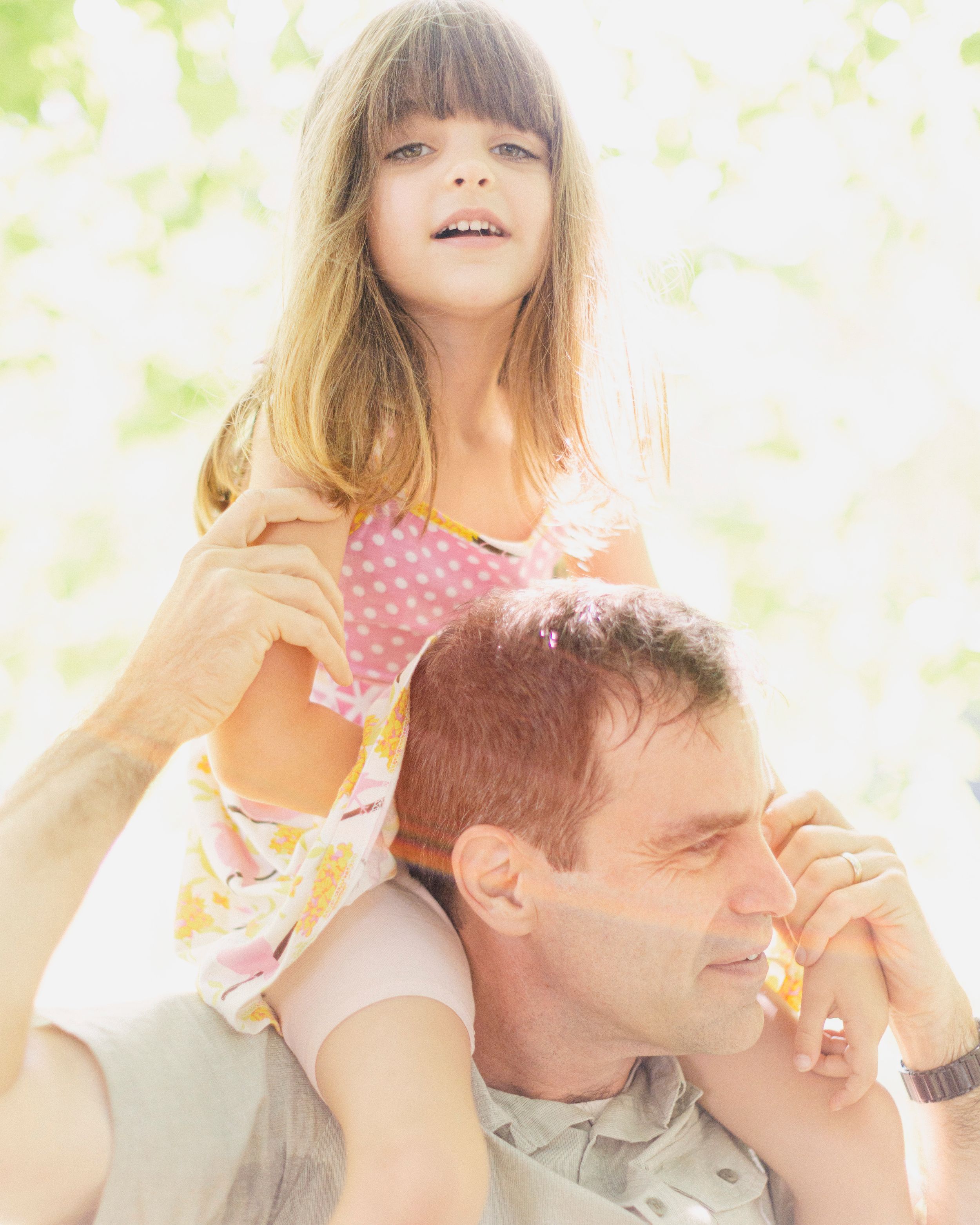 A child sits on shoulders in a sunny outdoor setting with soft bokeh background and warm lighting.
