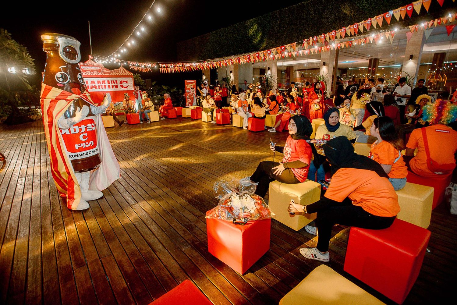 Colorful nighttime performance with traditional dancers and audience seated on red and yellow cushions on wooden deck.