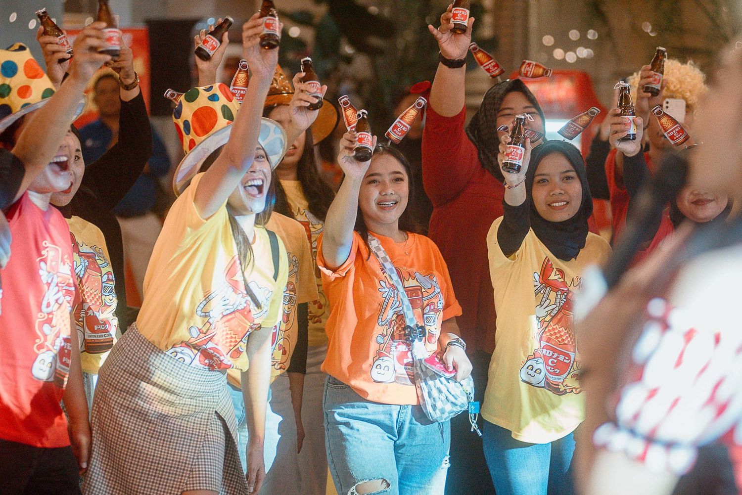 A crowd of enthusiastic people wearing colorful t-shirts raise their hands in celebration at an indoor event.