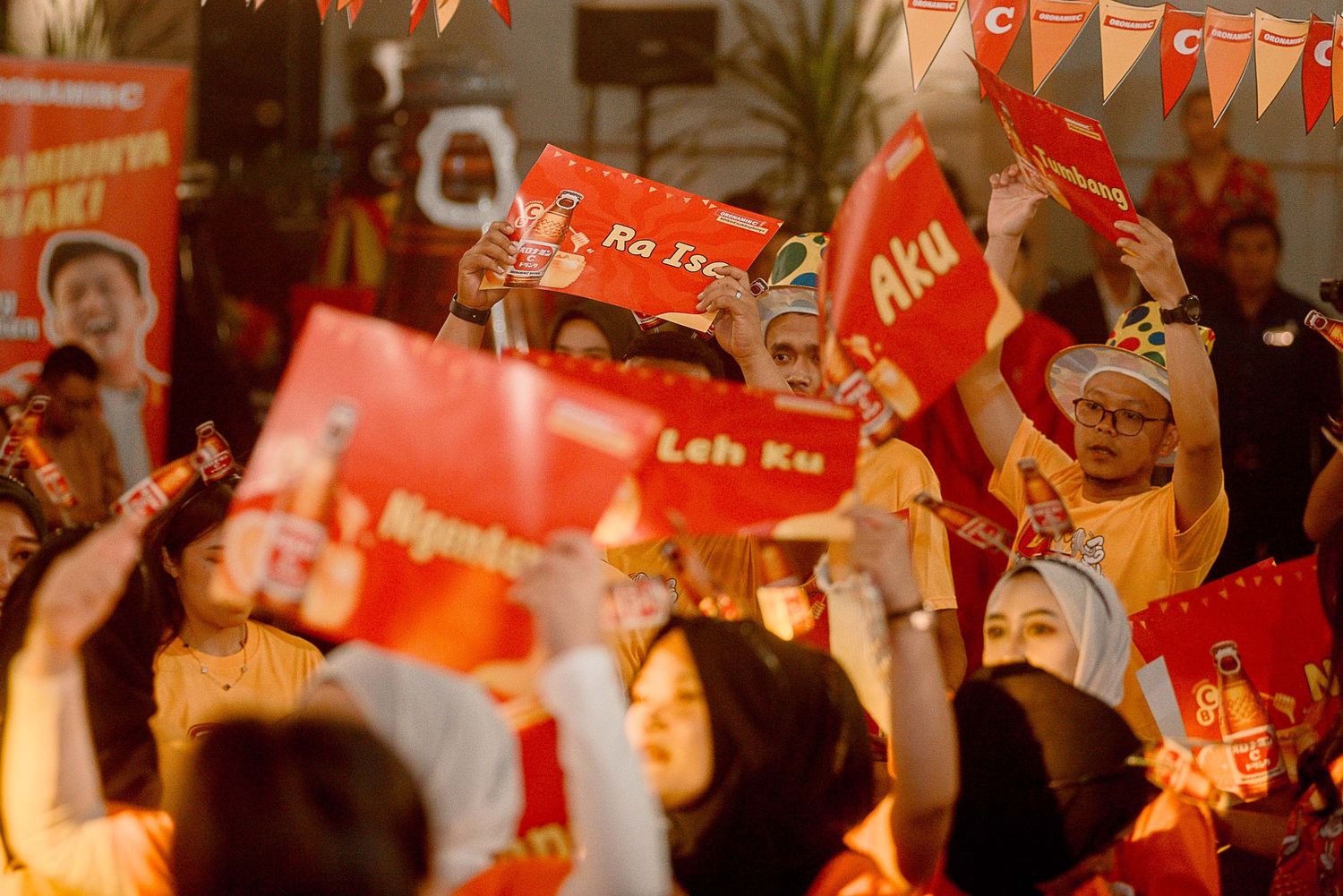 A crowd waves red flags and banners at a political rally with orange-tinted lighting and festive bunting overhead.