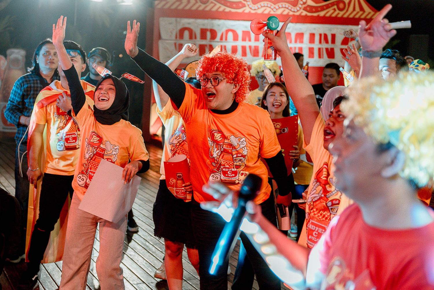 A crowd of people in orange shirts dancing and celebrating with raised hands at a colorful indoor event.