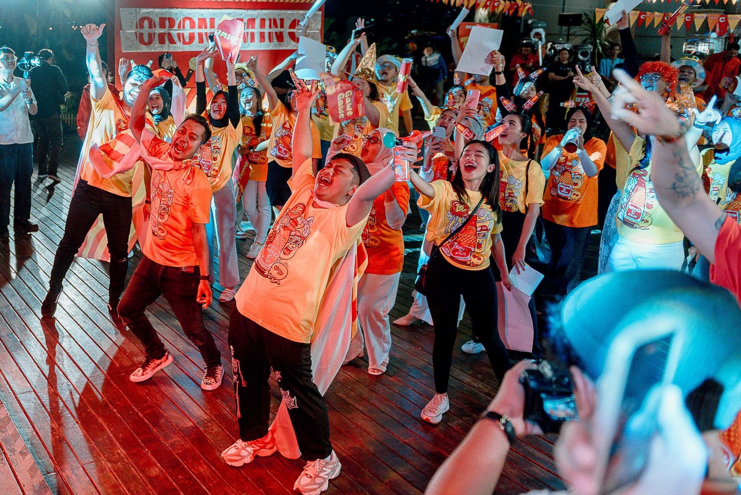 A group of dancers in orange shirts perform synchronized moves on a dance floor under colorful lighting.